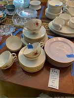 Stacked Lenox Roselyn china cups, saucers, and plates with pink rose design and gold trim, arranged on wood table with other clear and white glassware around