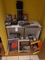 Photo showing CDs, cassette cases, and vinyl records arranged on a white shelf against a yellow wall.