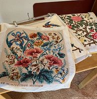 Overall view of three embroidered tapestry pieces stacked on a wooden surface showing floral designs and mesh canvas.