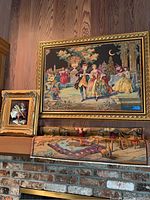 Framed tapestry depicting 18th-century style elegantly dressed figures in a garden setting mounted on a wooden paneled wall.