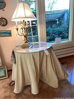 Vintage lamp with candelabra arms and white fabric shade sitting on round glass top table with beige fabric skirt, near window