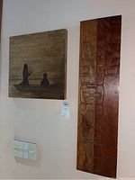 Photo showing the square canvas painting and the vertical wood carving side by side on a light wall, with a light switch below.