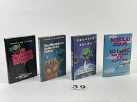 Four Douglas Adams sci-fi books standing upright, displayed with front covers visible: The Restaurant at the End of the Universe, The Hitchhiker's Guide to the Galaxy, Life, The Universe and Everything, So Long, and Thanks for All the Fish.