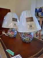 Two bedside lamps with floral ceramic bases and white linen shades still in plastic wrapping, shown on a tabletop.