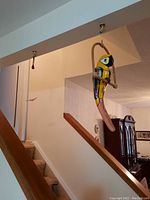 Wooden painted parrot hanging on a perch, displayed indoors by staircase.