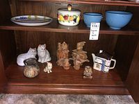 Shelf containing ceramic cat figurines, small animal figures, bark-style cat figurines, ceramic house creamer, and two blue mixing bowls