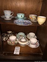 Front view of multiple fine bone china teacups and saucers arranged on wooden shelves, including a lidded sugar bowl.