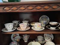 Shelf with seven fine bone china teacups and matching saucers arranged in two rows, displaying varied floral patterns and gold trimming.