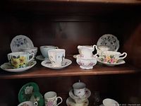 Display of seven teacups with matching saucers in wood shelf, includes floral patterns and white china creamer and sugar bowl with floral motif.