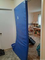 Side view of tall, rectangular blue medical mattress standing upright showing heavy, thick profile and blue vinyl cover with visible stitching