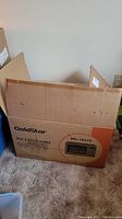 Full view of the GoldStar microwave packaging box placed on carpet next to a wall outlet, showing the brand and model number.