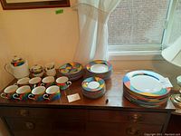 Photo showing multiple dishes including teacups with saucers, side plates, dinner plates arranged on a wooden dresser near a window with natural light