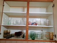 Front view of cabinet with shelves holding multiple types of vintage glassware: clear etched starburst, amber textured, ruby red stemmed glasses, green and white starburst frost glasses, smoky water and whisky glasses, and more.