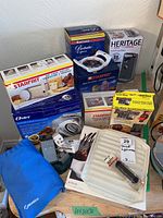 Lot overview showing boxed small appliances and accessories stacked including Oster hand mixers, Starfrit cheese maker, potato chippers, apple peeler, and furniture feet.