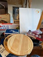 Lot of kitchen appliances and accessories including wooden cutting boards, boxes of kitchen appliances, and small kitchenware items arranged on a table.