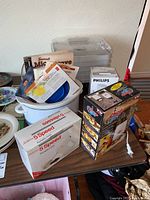 Overview of all items in the lot arranged on a table including boxes for dehydrator, garment steamer, food chopper, mixer, foldable colander and Crock Pot appliance.