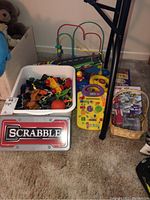Photo showing a white bin filled with various toy vehicles and toys, a large Scrabble tin, a yellow electronic educational toy, and a wire bead maze standing behind.