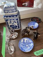 Photo showing tall Chinese ceramic container in blue and white with square shape and lid; two Wedgwood blue and white plates with floral and figurative designs; two blown glass swans; small animal figurines including elephant with damaged tusk and bull; stained glass hanging decoration.