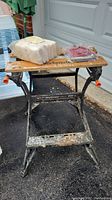 Side angle photograph of the Black & Decker Workmate portable workbench showing wooden top, metal frame with some rust and wear, and items placed on top: bags containing hardware hooks and stain applicators.