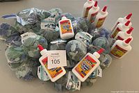 Photo showing 25 packs of mesh bags filled with blue, teal and white sea glass pieces, alongside 10 white squeeze bottles of Cra-Z-Art washable school glue with red caps.