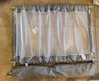 Folded gray fabric baby/pet gate laid flat with mesh panel and gray fabric edges, pictured with folded carrying case below it.