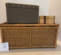 Large wicker basket chest with hinged lid, medium dark gray box with rivets, and small wooden decorative box with bow design arranged for size comparison.