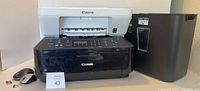 Two Canon Pixma printers in white and black with no visible power cords next to black shredder receptacle can.