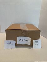 Photo showing a single assembled cardboard box sealed with packing tape, with handwritten labels indicating quantity (25) and box size (12x12x6 inches)