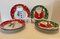 Photo showing 8 round ceramic plates stacked in two piles, 4 displaying a snowman design with red background and white polka dots, and 4 with a Santa Claus design on a green background with white polka dots.