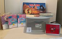 Monopoly Utah Edition board game box, Sterilite plastic drawer unit, two books, and Babybel metal lunch box arranged on table.