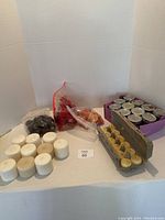 Wide shot showing assortment of wax candles including white pillar candles, dark colored candles in bags, and boxed tealight candles.