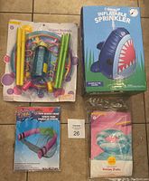 Overhead photo showing the bubble wand set in original packaging, the inflatable sprinkler box, the swim tube packaged in plastic, and the pool noodle chair accessory in package.