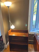Photo showing wood book shelf with two shelves and raised sides next to a floor lamp with two light fixtures on a hardwood floor.