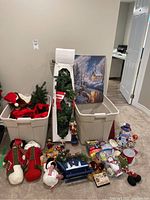 Christmas decorations including small artificial tree, winter painting, assorted ornaments, string lights, and figurines in storage bins and arranged on floor