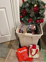 Plastic bin with assorted Christmas wrapping paper, bows, ribbons, and two wreaths resting on the bin. A reusable red gift bag and cloth gift bags are visible at the front of the bin.