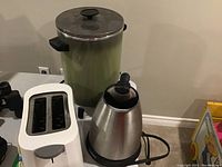 All three appliances shown: large green percolator coffee pot with lid missing knob, stainless steel Cuisinart kettle, and white Black & Decker toaster.