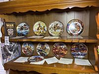 Eight Dominion China Ltd wartime collector plates displayed on a wooden hutch shelf with certificates and one plate laying in front.