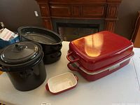 Full view of all cookware items on table, including red covered pan with lid, small microwave dish, and black roaster and steamer.