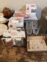 Photo showing multiple boxed and unboxed glassware items including crystal glasses, Christmas-themed glasses, and set of cut glass salt cellars with spoons on wooden surface.