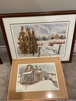Two framed paintings shown with one in the foreground and one leaning behind, both set on a floor against wall
