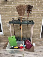Full view of garden stand holding various garden tools including broom, shovel, hand tools, and red watering can to the side with a plastic bin containing smaller hand tools and a kneeling pad.