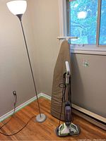 Photo of the floor lamp, metal ironing board leaning against the wall, and Bissell floor finishing machine upright beside them.