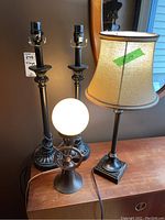 Four lamps on wooden surface against wall including two tall black metal candlestick-style lamps without shades, one lamp with beige shade, one small lamp with white globe and gear base.