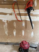 Overall view of lot showing black and decker electric leaf blower hanging on wall, orange hand bow saw hanging next to it, and red shop vac on floor.