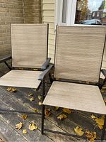 Two outdoor folding chairs with metal frames and beige fabric seats and backs shown side by side on wooden deck with autumn leaves.