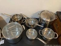 Overview of seven stainless steel pots and pans including stockpots, saucepans, and a strainer pot with some lids present.