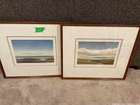 Both framed landscape prints side by side on carpeted floor against wall.