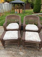 Two brown wicker chairs and two matching footstools on outdoor wooden deck, with cushions featuring leaf pattern.