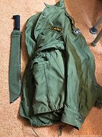 Photo showing the olive green vintage U.S. Army jacket laid flat with a machete in a green sheath alongside it.