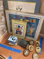 Nine art pieces including framed art and small oval frames arranged on carpet with blue tape lines visible. Included is a box of glass photo coasters and a stack of plastic picture frames.